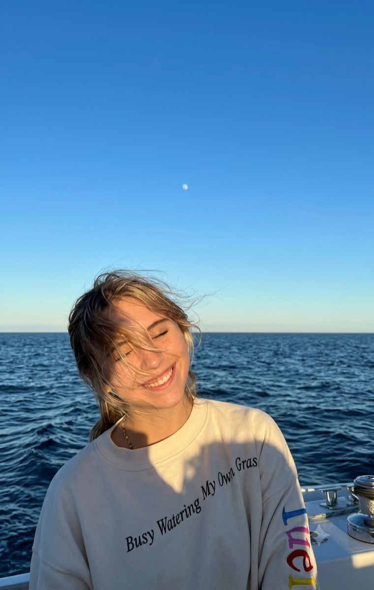 Camille Devaney smiling with her eyes closed and wind in her hair, on a boat at sea during golden hour, the deep blue ocean and a clear sky behind her.